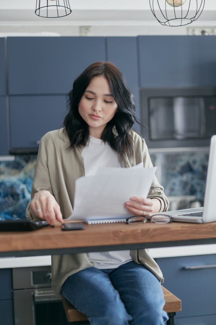 about-us-01 A young woman reviews documents and uses her laptop in a modern kitchen setting.
